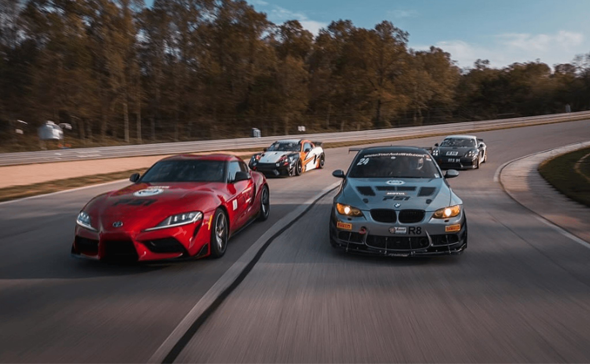 Four sports cars race down a curving track, surrounded by trees. A red car leads, conveying speed and competition under a clear blue sky.